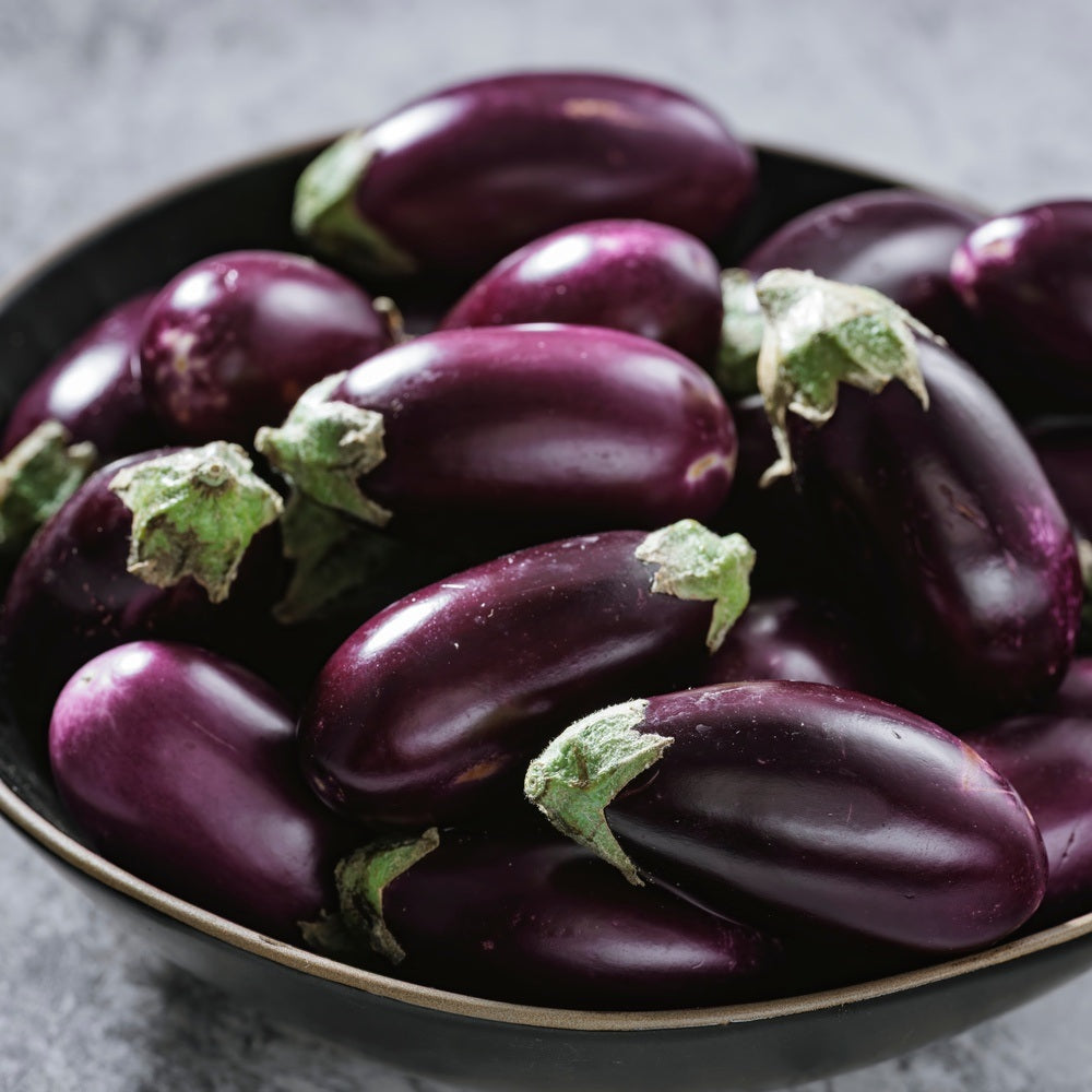 Small oval eggplants with glossy deep-purple skin and pale green stem caps, piled together in a black bowl.