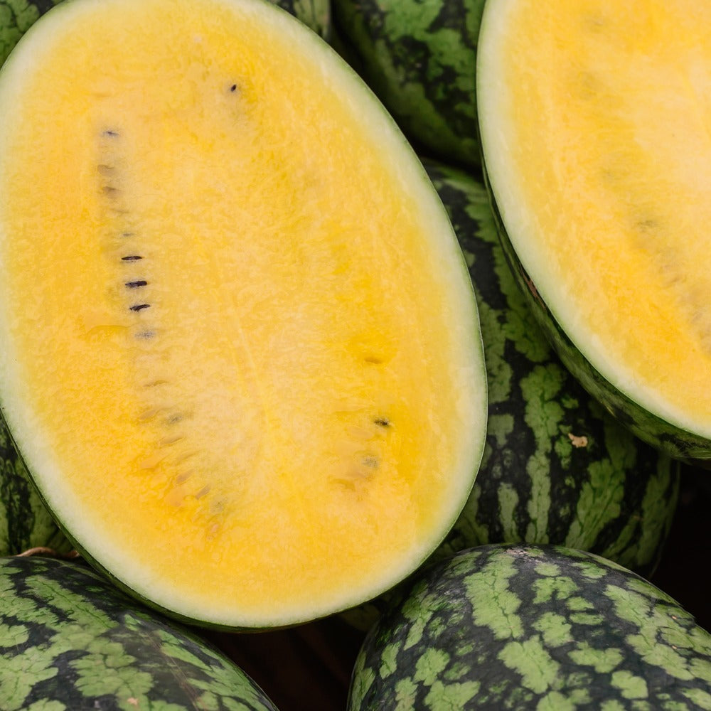 Halved watermelon revealing bright yellow flesh with a central line of dark seeds, surrounded by whole dark green striped watermelons.