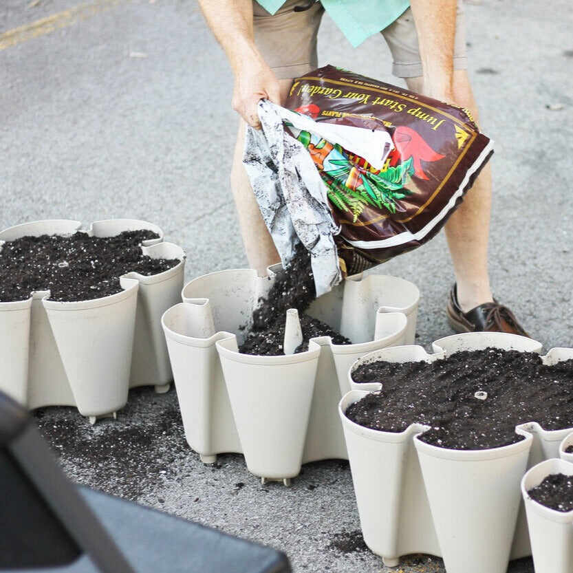 A person pours dark potting soil from a labelled garden soil bag into the scalloped pockets of a stacked, tan-coloured GreenStalk vertical planter set on a paved driveway.