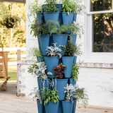 A five-tier blue GreenStalk vertical planter on a porch, each tier's pockets filled with trailing herbs and foliage including dusty miller, coleus, and fennel, set against a white brick wall.