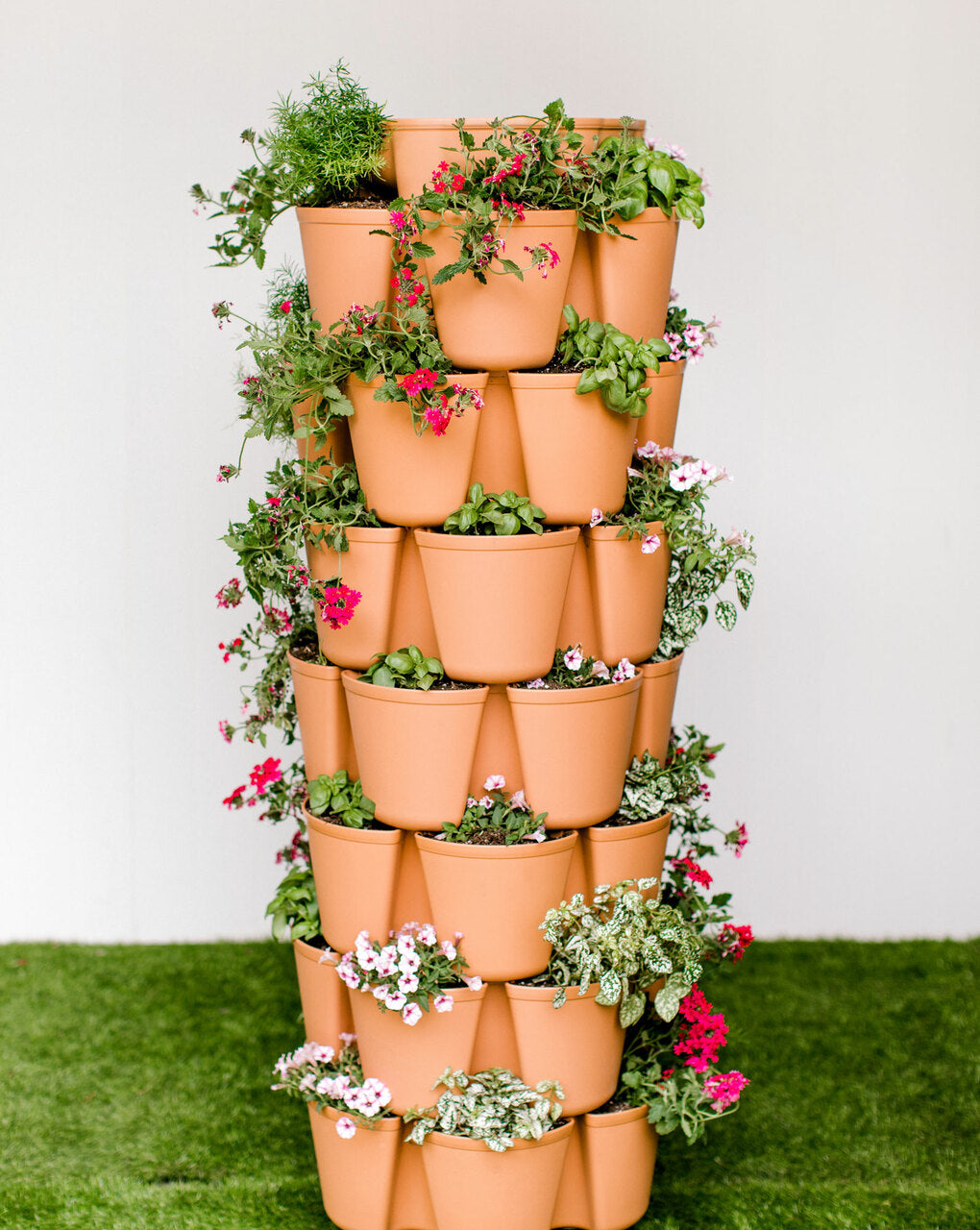 Terracotta-colored seven-tier vertical planter tower with staggered plastic pots on each level, densely planted with trailing red verbena, white petunias, basil and variegated foliage, standing on artificial turf against a plain wall.