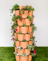 Terracotta-colored seven-tier vertical planter tower with staggered plastic pots on each level, densely planted with trailing red verbena, white petunias, basil and variegated foliage, standing on artificial turf against a plain wall.