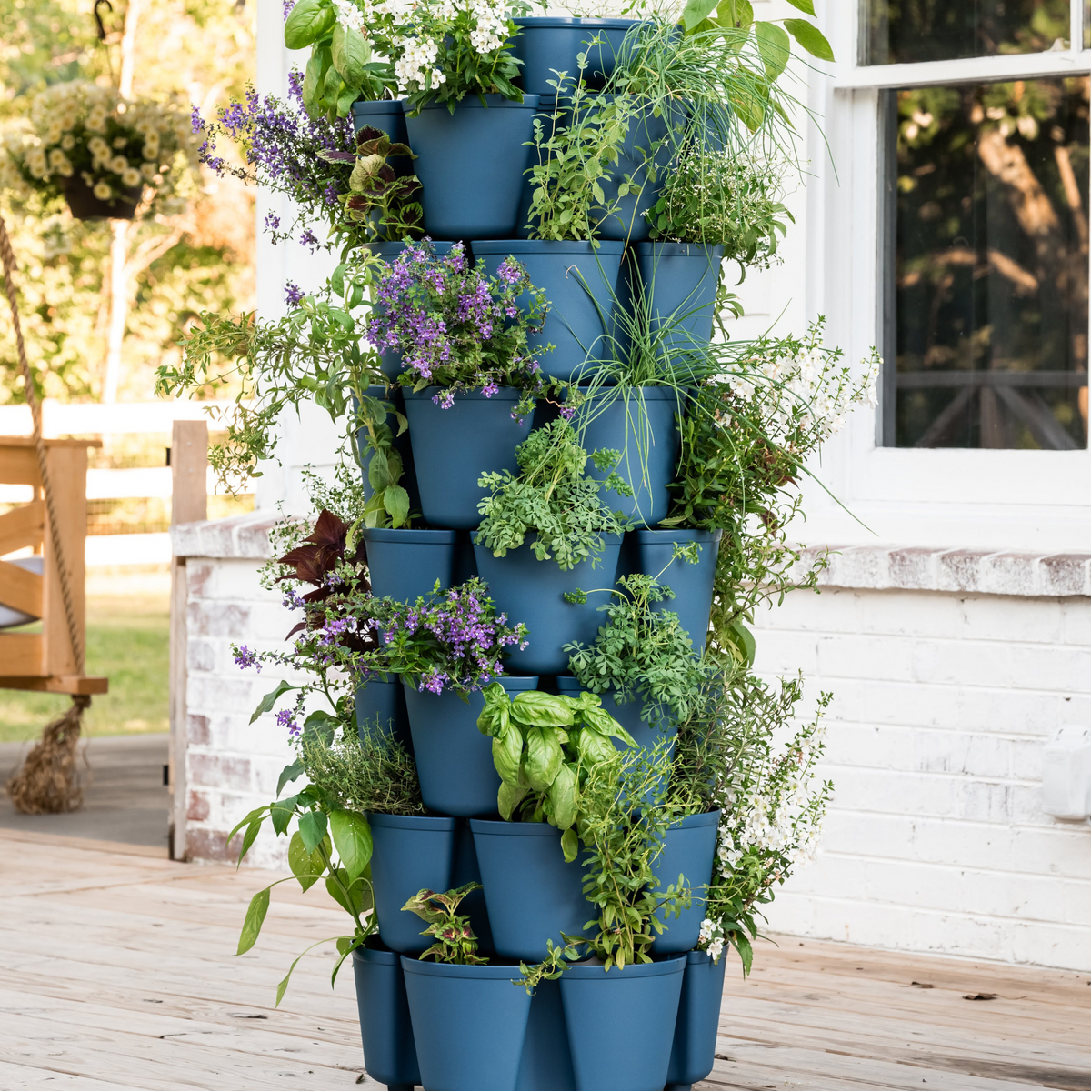 A seven-tier blue GreenStalk vertical planter overflowing with basil, oregano, chives and trailing purple-flowered herbs, standing on a wooden porch beside a white brick wall and window.