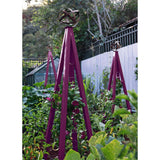Purple pyramid-shaped metal Akoris Garden Obelisk Large trellises with bronze sphere finials set among lush green foliage and purple flowering plants