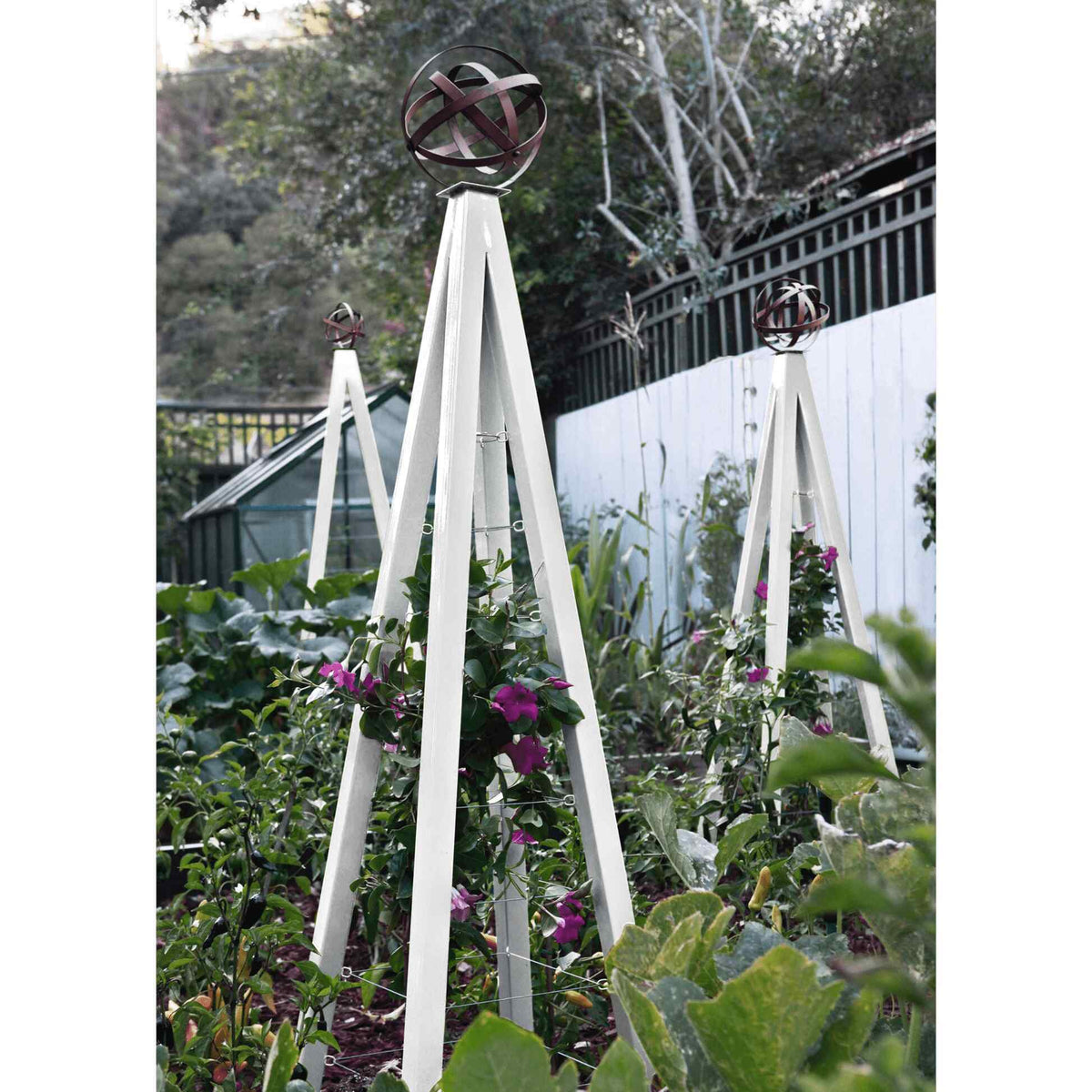 White pyramid-shaped metal Akoris Garden Obelisk Large trellises with bronze sphere finials set among lush green foliage and purple flowering plants