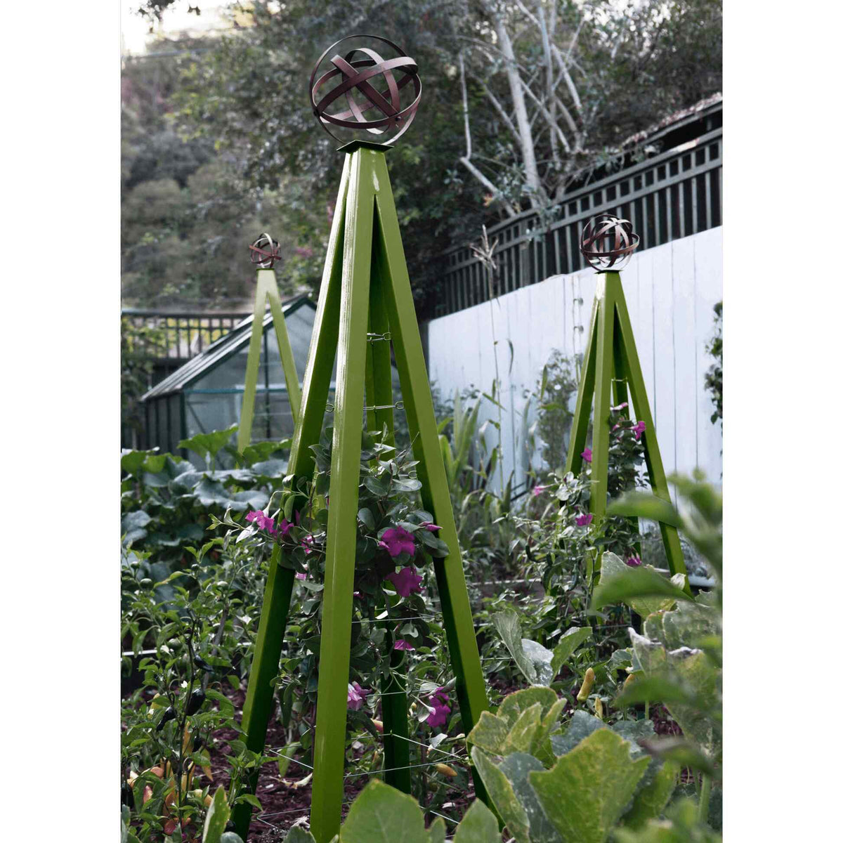 Olive green pyramid-shaped metal Akoris Garden Obelisk Large trellises with bronze sphere finials set among lush green foliage and purple flowering plants