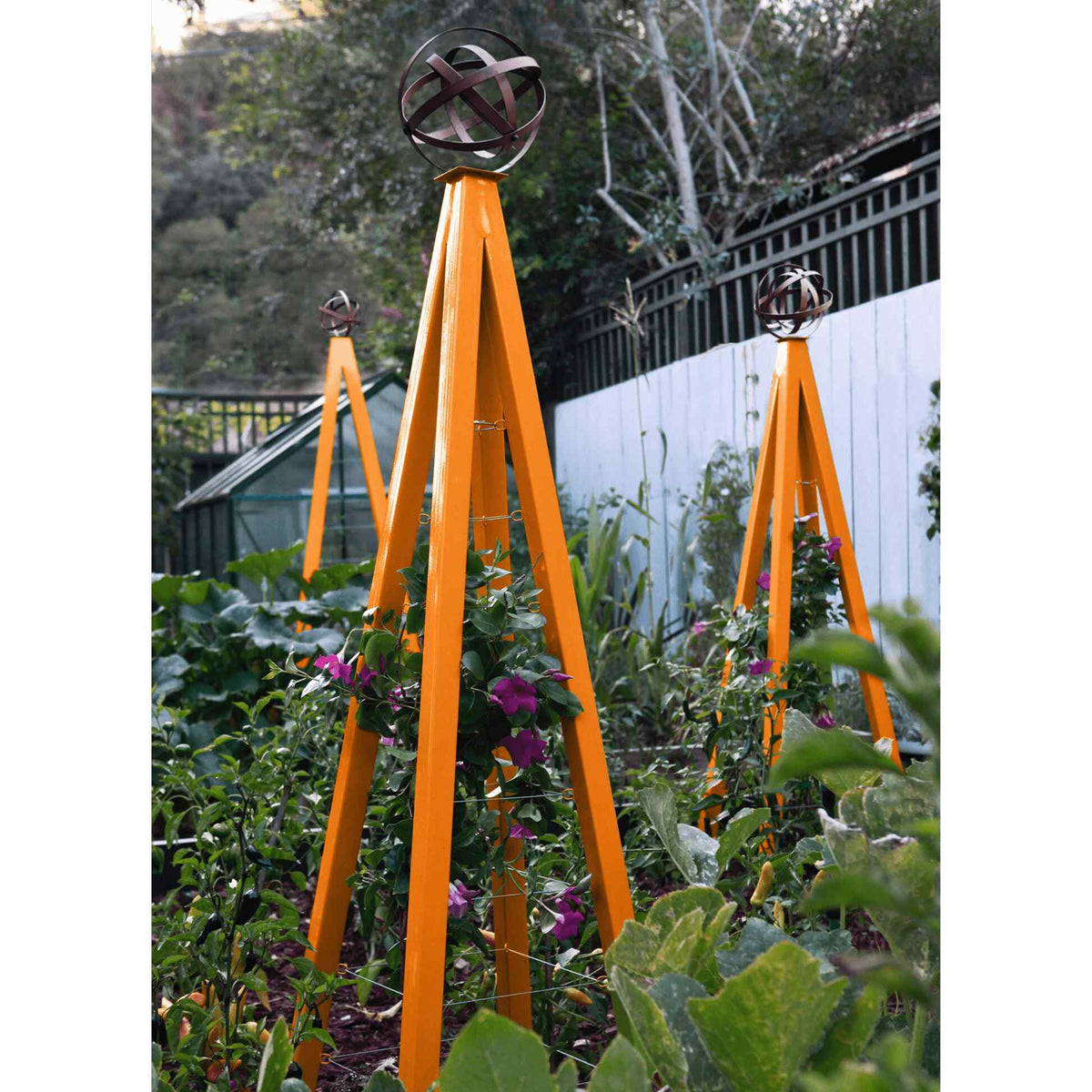 Orange pyramid-shaped metal Akoris Garden Obelisk Large trellises with bronze sphere finials set among lush green foliage and purple flowering plants