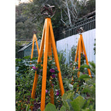 Orange pyramid-shaped metal Akoris Garden Obelisk Large trellises with bronze sphere finials set among lush green foliage and purple flowering plants