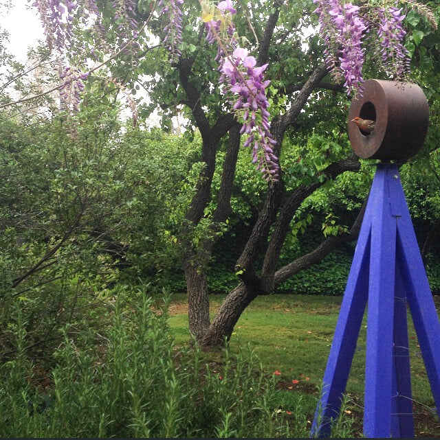 Rust-finished A-frame Bird Cafe with Trellis topped with a circular bird feeder, surrounded by green shrubs and orange flowering vines