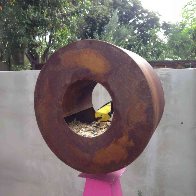 Close-up view of a yellow bird inside a rust-finished circular steel bird feeder against a light concrete wall