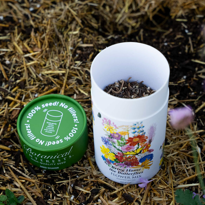 Open Botanical Interests Bring Home the Butterflies seed shaker tube next to its green lid on straw mulch.
