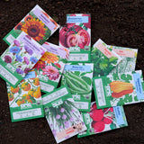 An assortment of colorful Epic Gardening seed packets—including flowers, herbs, and vegetables like sunflowers, tomatoes, beans, and radishes—scattered across dark, rich garden soil.