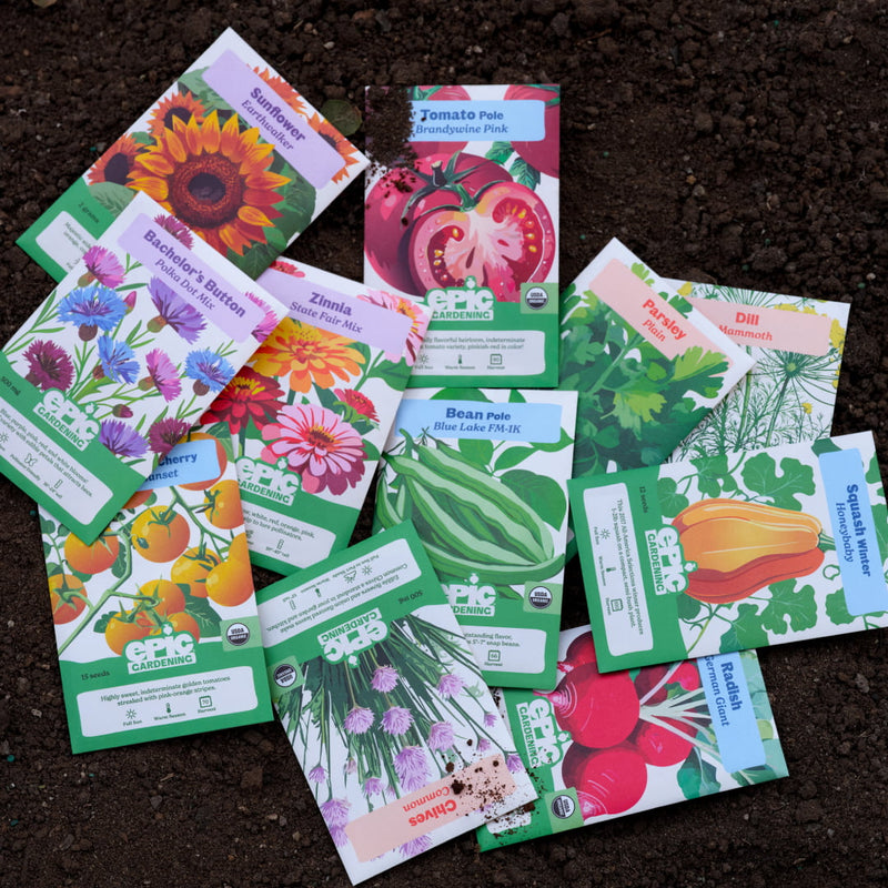 An assortment of colorful Epic Gardening seed packets—including flowers, herbs, and vegetables like sunflowers, tomatoes, beans, and radishes—scattered across dark, rich garden soil.