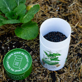 An open Botanical Interests Common Buckwheat seed shaker container filled with seeds resting next to its green lid on a straw mulch background.