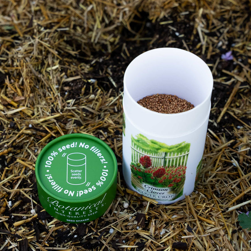 An open Botanical Interests Crimson Clover Seed Shaker container filled with seeds resting next to its green lid on a straw mulch background.