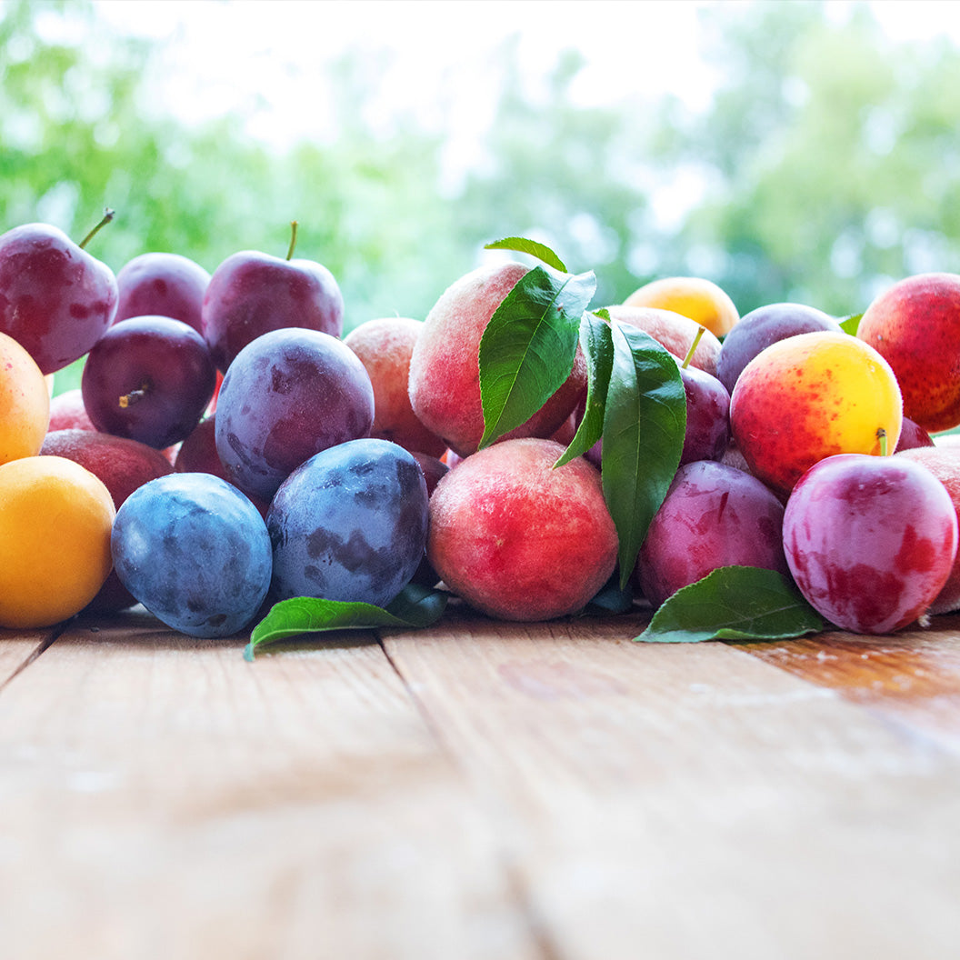 Assortment of fresh stone fruits—including purple plums, yellow apricots, and pinkish-red peaches—arranged on a light wooden surface against a blurred green background.