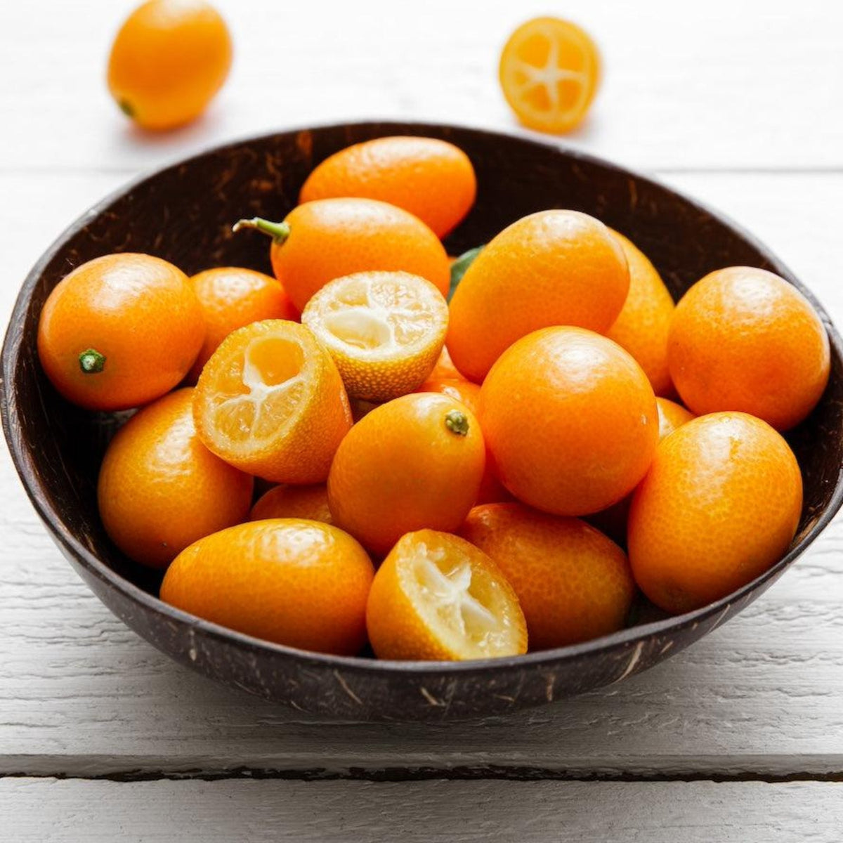 A dark wooden bowl filled with fresh, bright orange Fukushu kumquats, including whole fruits and cut halves showing juicy pulp, resting on a white wooden surface.