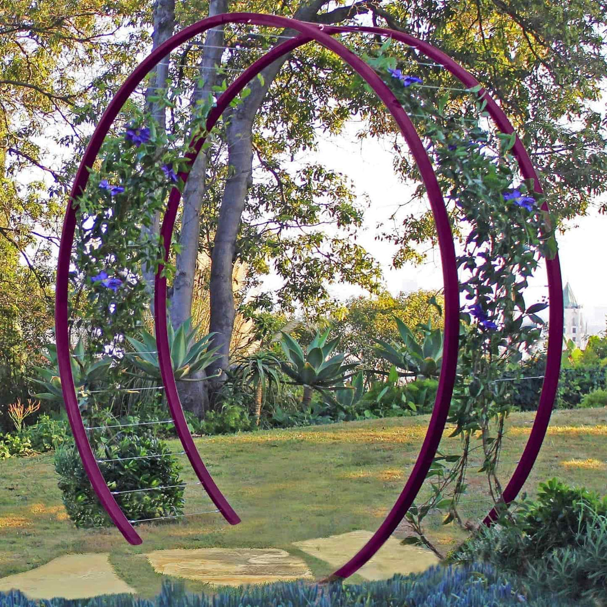 Purple circular metal Gracie Modern Arbor forming an arched gateway over a grassy path in a garden with trees and foliage