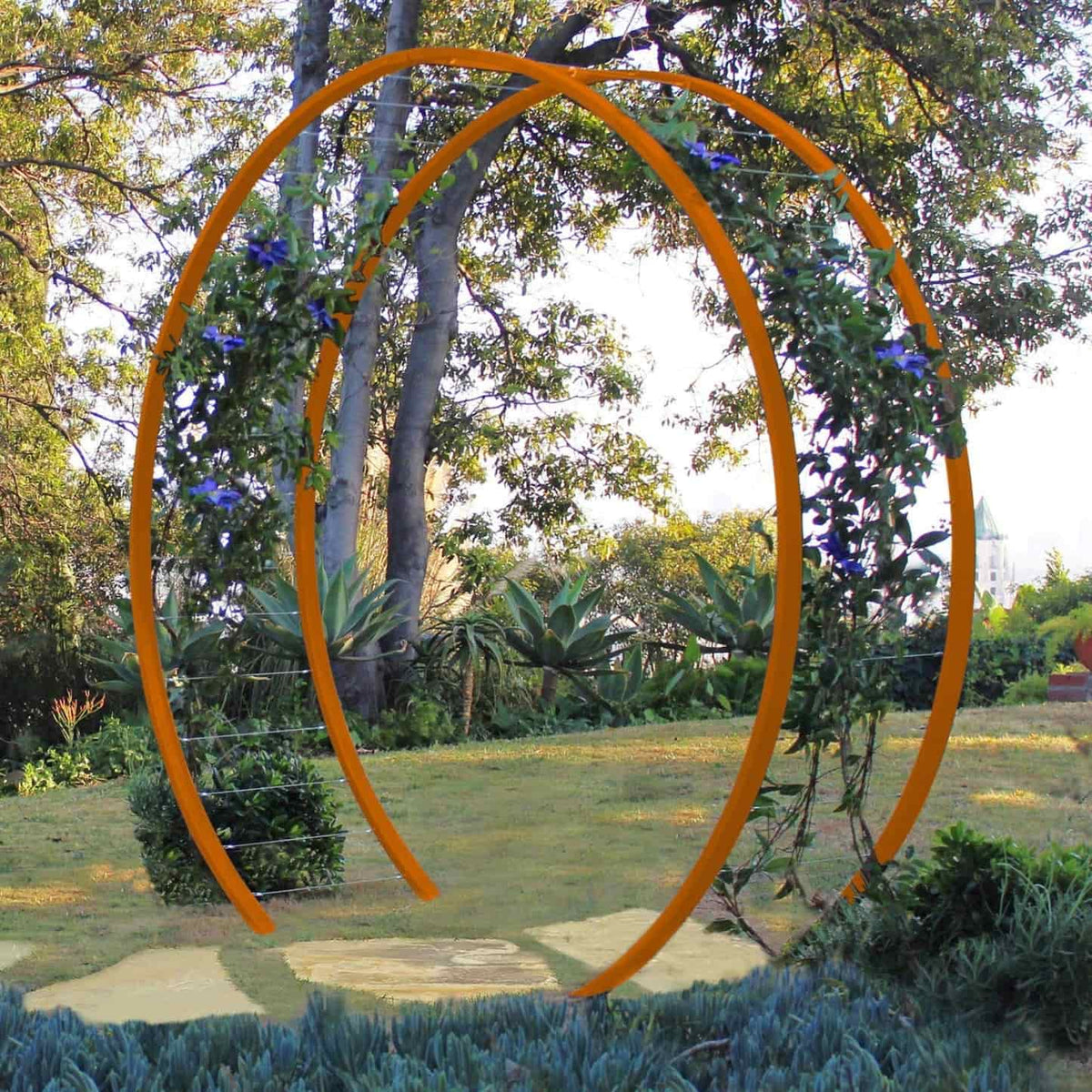 Bright orange circular metal Gracie Modern Arbor forming an arched gateway over a grassy path in a garden with trees and foliage