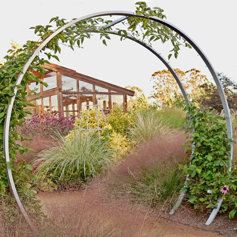 Silver circular metal Gracie Modern Arbor forming a garden archway over a path, with a wooden greenhouse in the background