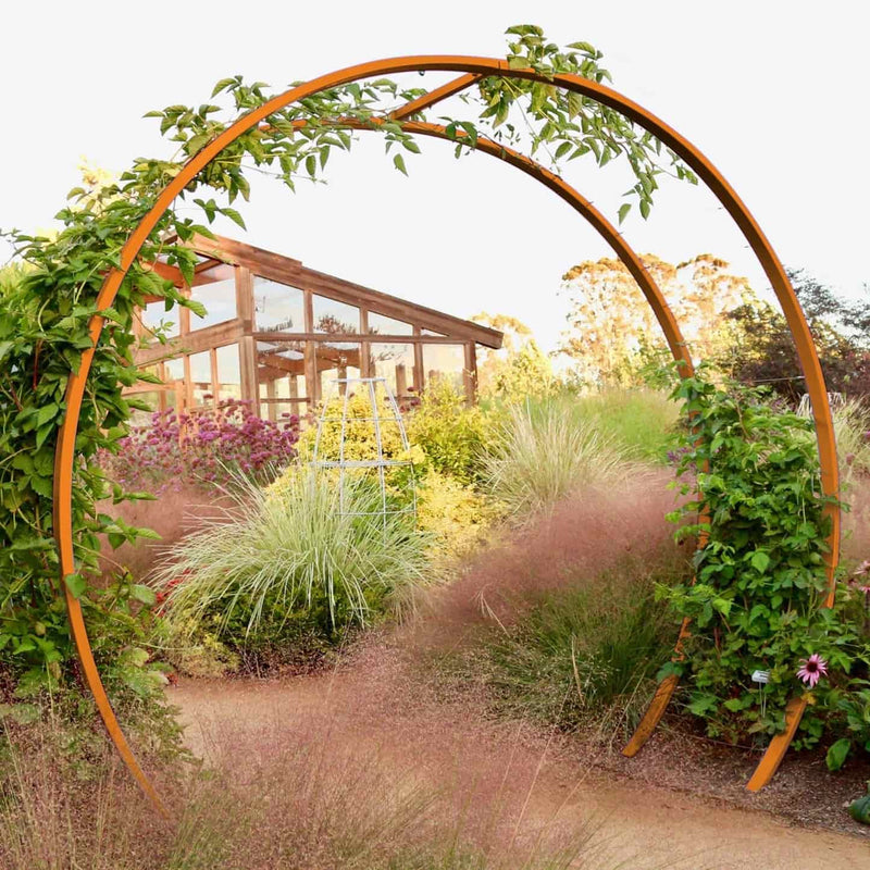 Rust-finished circular metal Gracie Modern Arbor forming an arched entryway over a gravel path in a lush garden setting