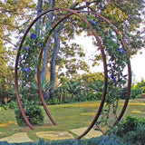Rust-finished metal circular Gracie Modern Arbor forming a garden entryway next to a mature tree and lawn