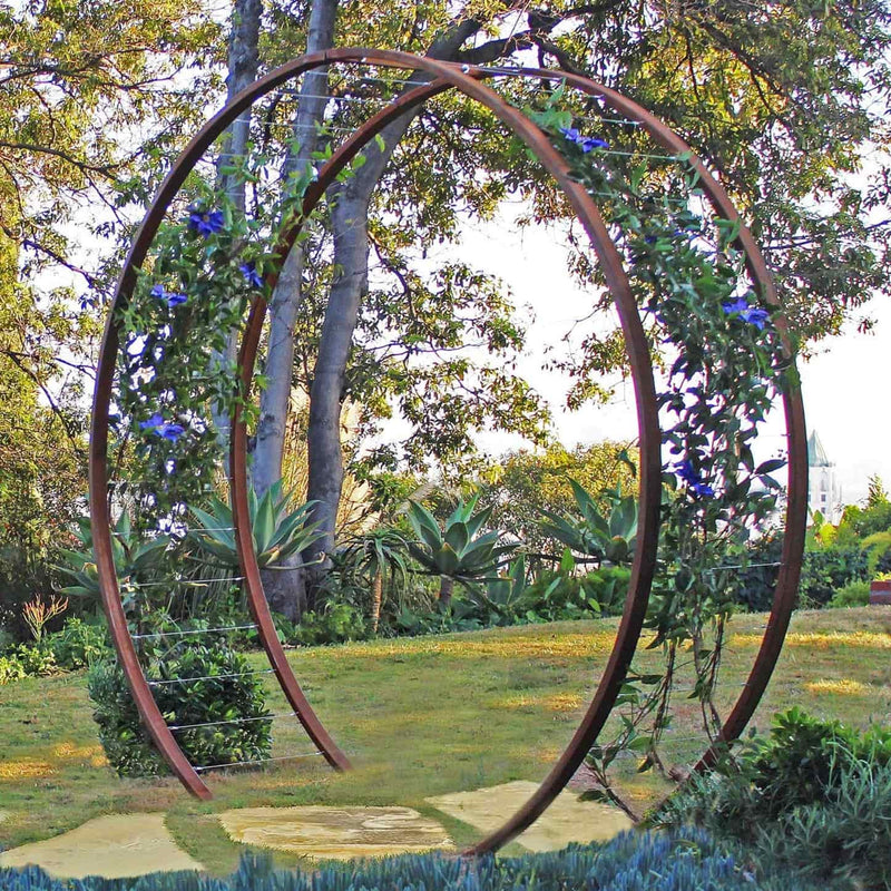 Rust-finished metal circular Gracie Modern Arbor forming a garden entryway next to a mature tree and lawn