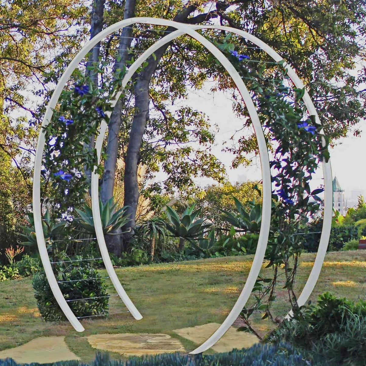 White circular metal Gracie Modern Arbor forming an arched gateway over a grassy path in a garden with trees and foliage