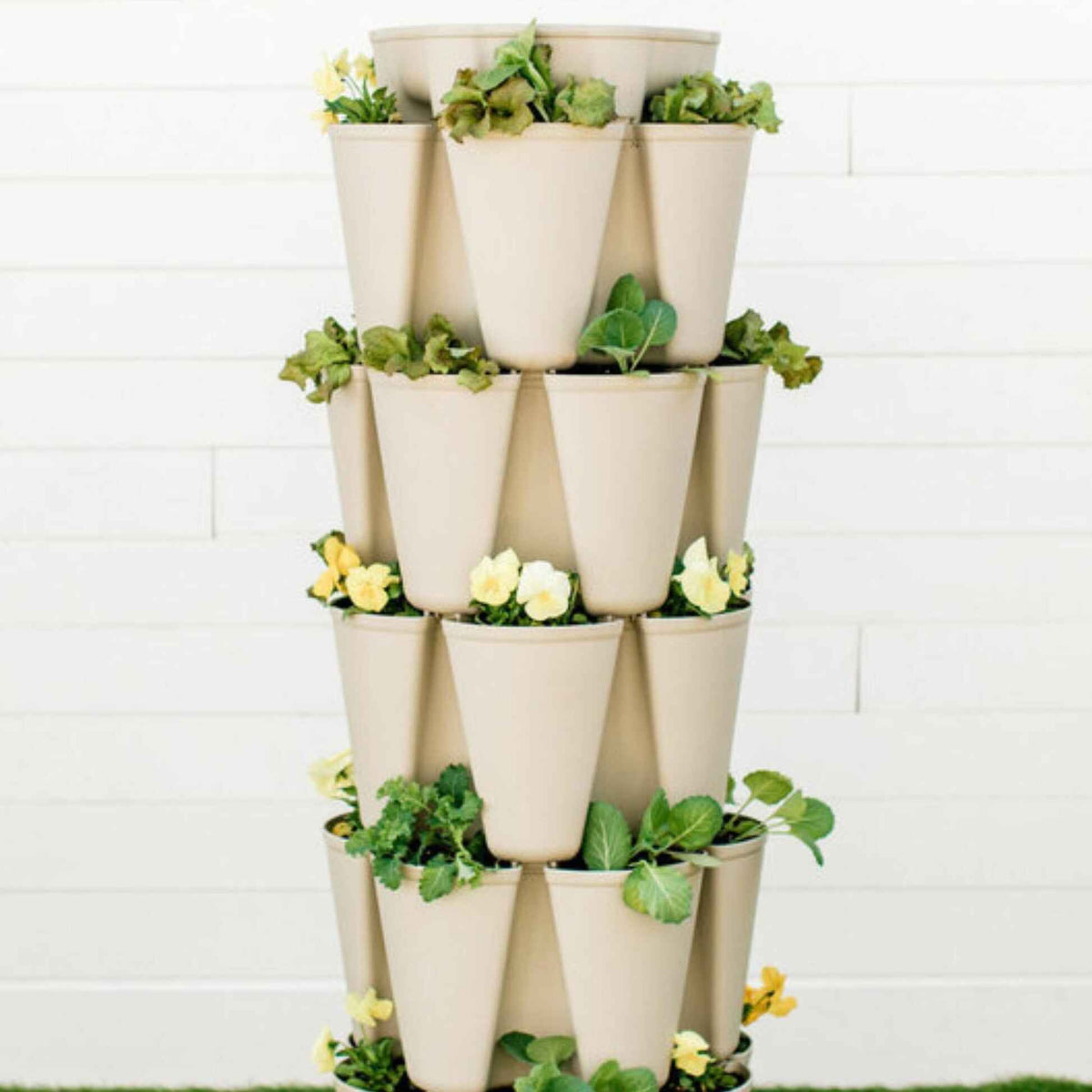 A stacked five-tier vertical planter with tan, cone-shaped pockets arranged around each level, planted with leafy greens and yellow pansies, standing against a white shiplap wall.