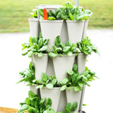 Stacked tiers of a grey GreenStalk vertical planter densely filled with leafy green seedlings, water droplets on the foliage, an orange plant marker tucked among the top leaves, set outdoors against a blurred lawn.