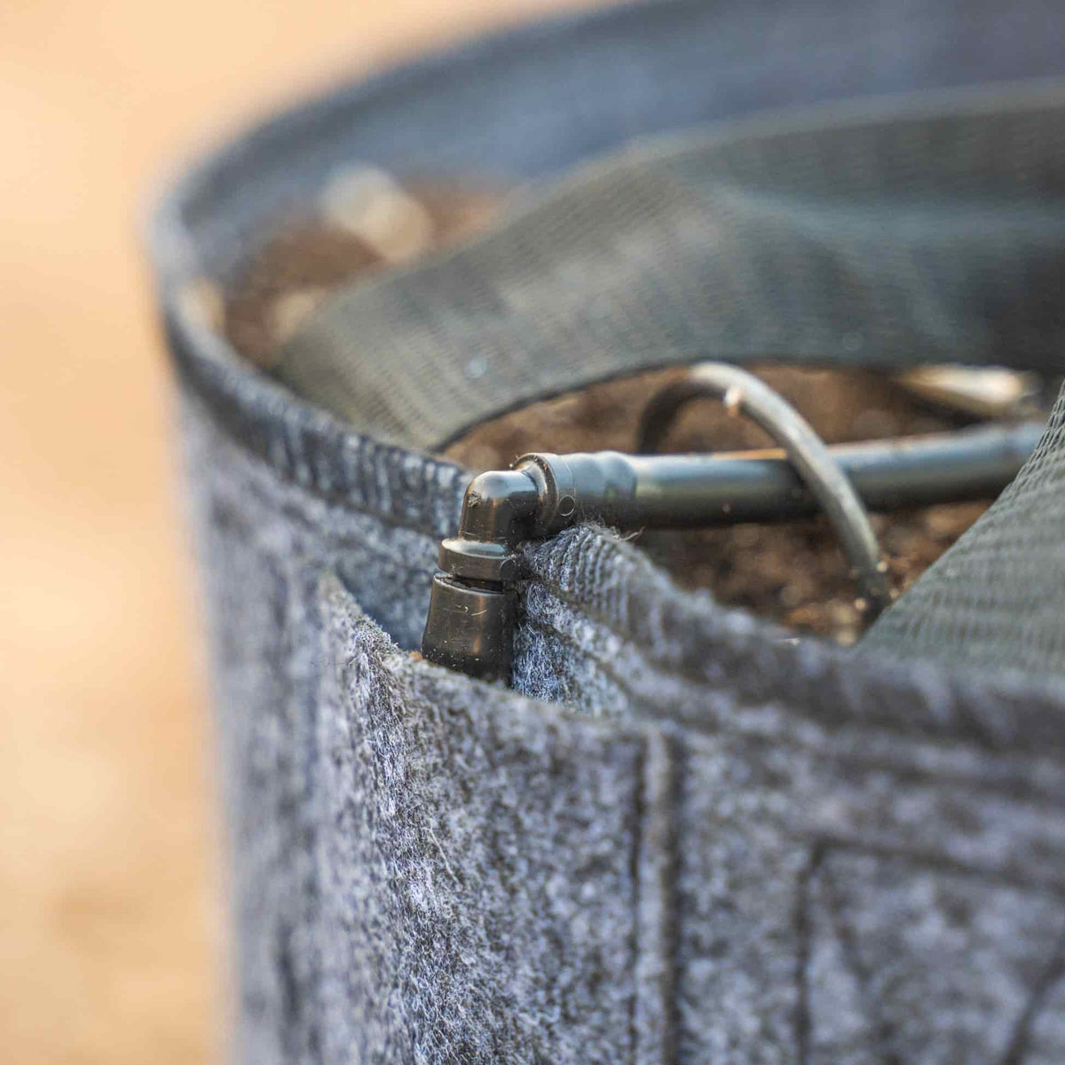 Close-up of a grey felt grow bag filled with soil, showing a small black irrigation fitting and curved drip tubing threaded over the rim.
