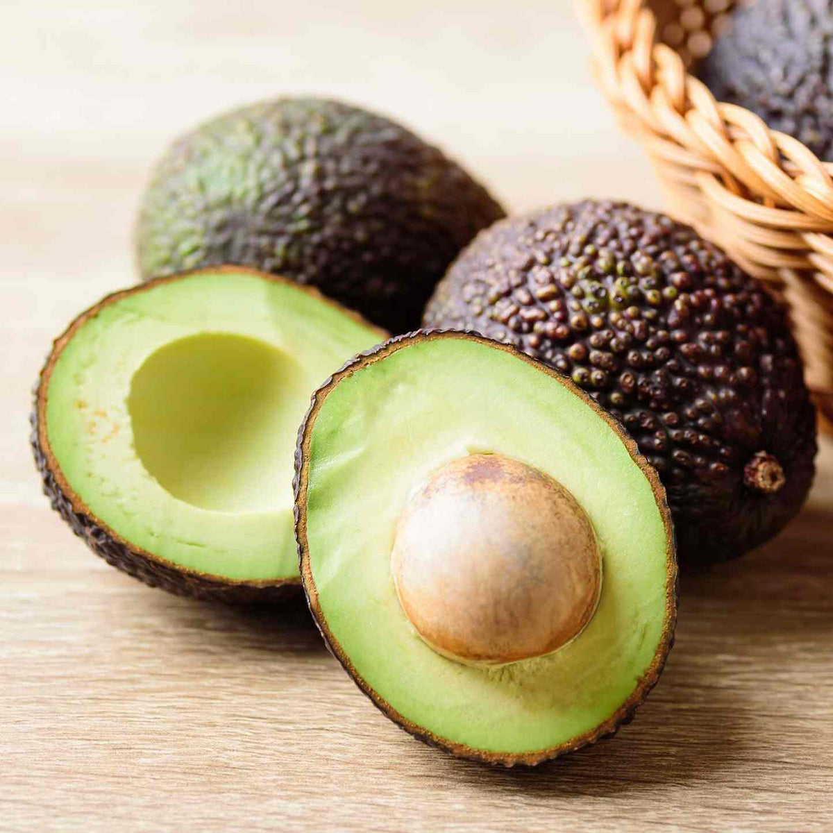 Halved Hass avocado with pit alongside a whole avocado on a wooden surface.