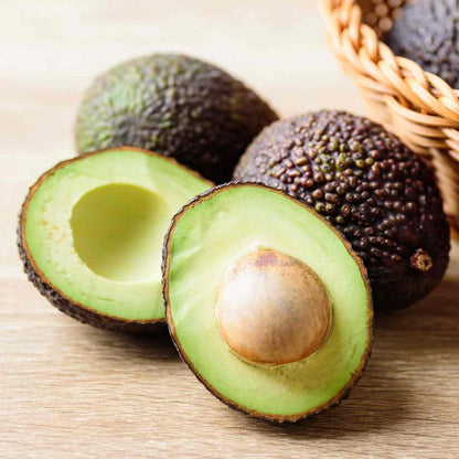 Halved Hass avocado with pit alongside a whole avocado on a wooden surface.