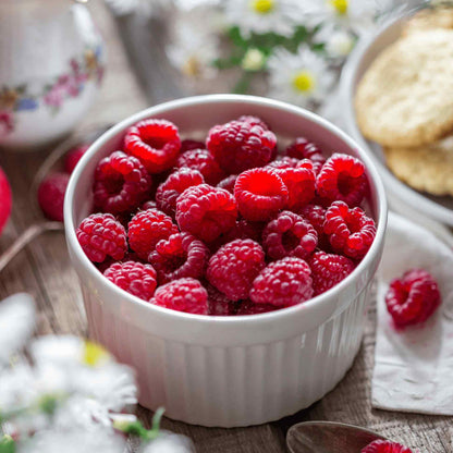 White ceramic bowl filled with freshly harvested red raspberries from a Heritage Raspberry plant, set on a rustic wooden table beside flowers and baked treats.