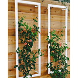 Two white metal rectangular Ina Wall Trellises mounted on a light wood wall with flowering vines climbing through their wire grids