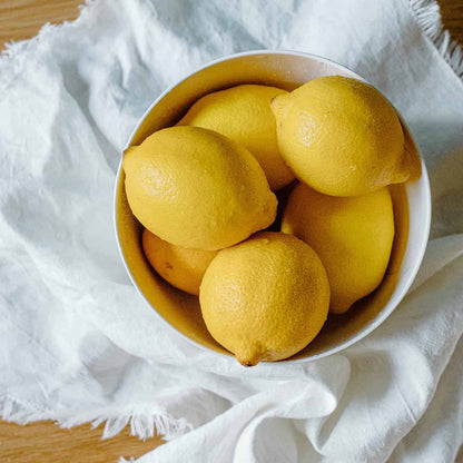 White bowl filled with fresh, bright yellow Improved Meyer Lemons resting on a textured white linen