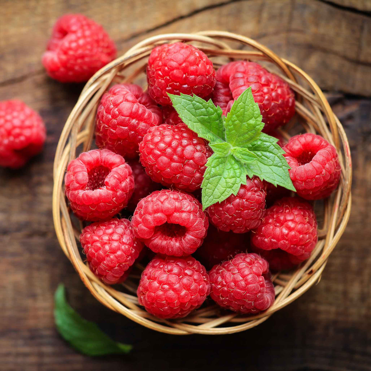 Top-down view of a woven basket filled with fresh, bright red raspberries topped with a sprig of fresh green mint, resting on a rustic wooden surface.