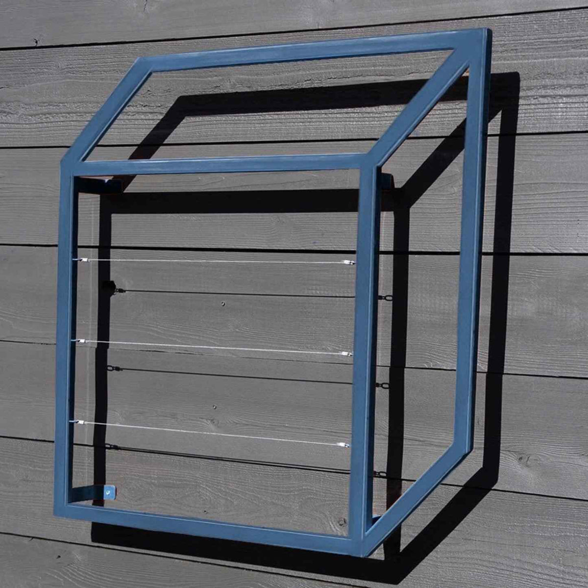 Blue cubic metal Pablo Wall Trellis mounted on a gray wooden panel wall, throwing strong geometric shadows