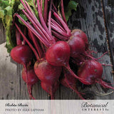 A freshly harvested bunch of Robin beets with deep red-purple bulbs, trailing roots, and magenta-veined leaf stems, laid on weathered wooden boards, labelled 