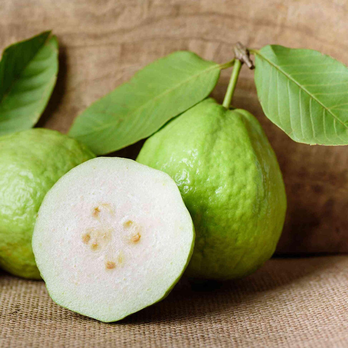 Whole light green guavas and a cut cross-section of a White Guava showing its pale flesh and seeds, set on a textured burlap background with green leaves.