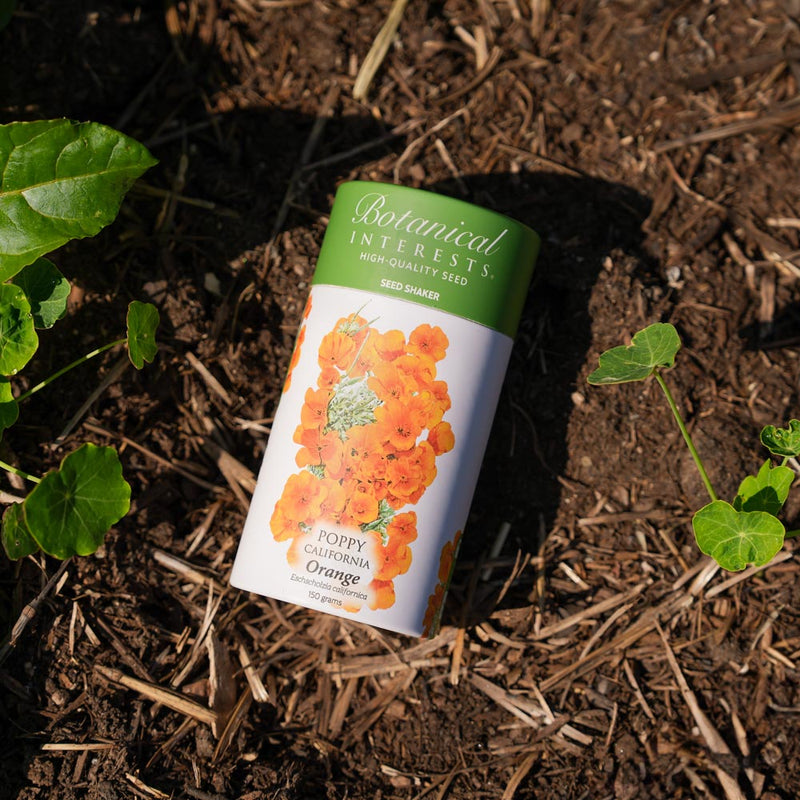 Botanical Interests Orange California Poppy Seed Shaker canister on soil with plants around