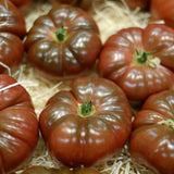 Close-up of ribbed Cherokee Carbon tomatoes with deep reddish-brown, glossy skins and green stems, nestled together in wood-shaving packing straw.