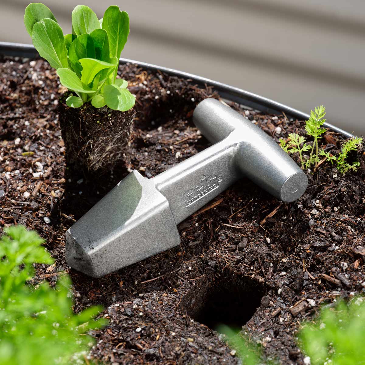 A metallic silver Epic Planting Dibble with a T-handle, resting on a soil with potted plants in the background.