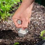 A close-up of a hand gripping the Epic Planting Dibble, pressing its square metal tip into dark, organic soil surrounded by green foliage.