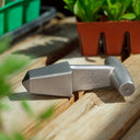 A metallic silver Epic Planting Dibble with a T-handle, resting on a wooden surface with potted plants in the background.