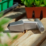 A metallic silver Epic Planting Dibble with a T-handle, resting on a wooden surface with potted plants in the background.