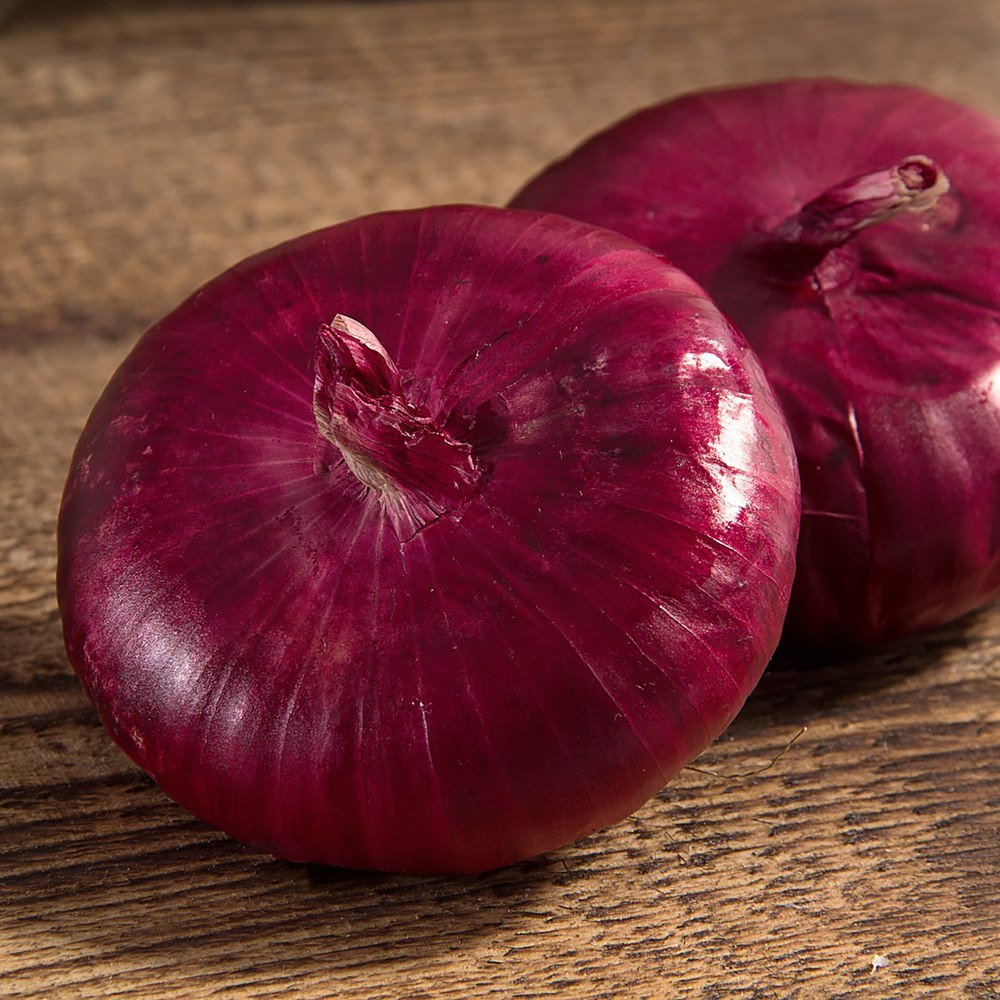 Two glossy, deep magenta-red onions with papery skin and dried root tufts, resting on a rustic wooden surface.