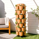 Terracotta-colored GreenStalk 7-tier vertical planter on wheeled casters, each tier ringed with cascading pots of leafy greens and white flowers, standing on artificial turf beside a wooden outdoor sofa and white shiplap wall.