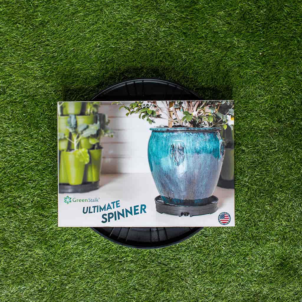Black round GreenStalk Ultimate Spinner base sitting on artificial turf, its packaging showing photos of a glazed blue-grey planter and a stacked green vertical garden tower, with a USA-made badge.