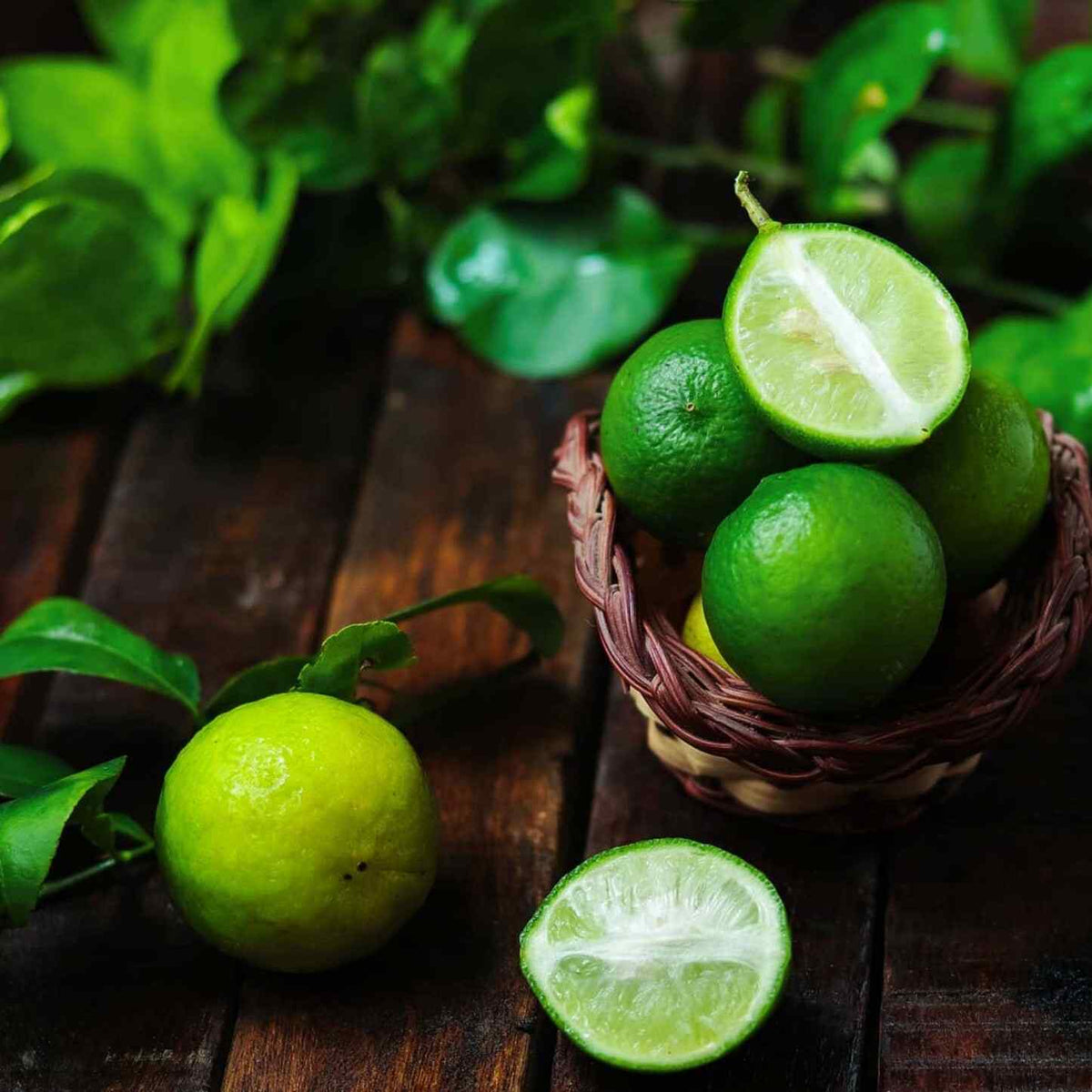 Whole and halved Key Limes in a woven basket on a rustic wood surface.